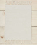 A small rectangular ivory Bath Mat with fringe on the short ends lies on a light wood floor with visible grain.