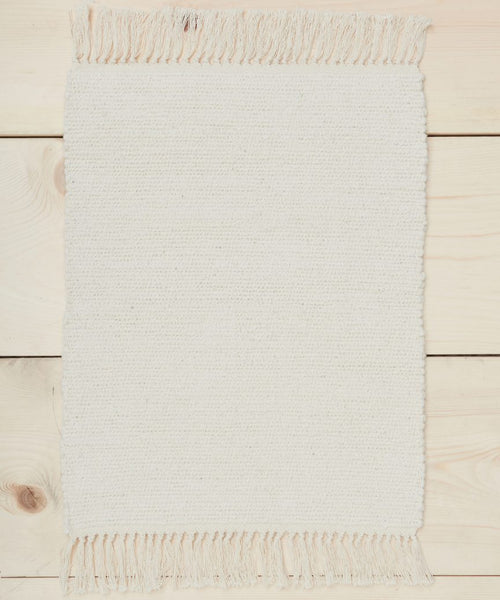 A small rectangular ivory Bath Mat with fringe on the short ends lies on a light wood floor with visible grain.