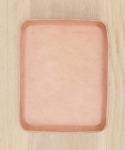 A light pink, square, shallow Leather Catchall with rounded edges rests on a light wood surface—an elegant accessory handmade in Los Angeles.