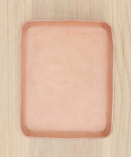 A light pink, square, shallow Leather Catchall with rounded edges rests on a light wood surface—an elegant accessory handmade in Los Angeles.