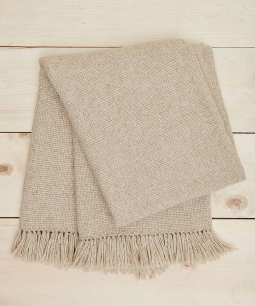 The Large Alpaca Basketweave Throw in beige, featuring super-soft alpaca and fringe edges, is neatly folded on a light wooden surface.