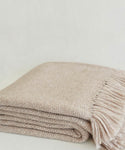 The Large Alpaca Basketweave Throw, featuring a soft woven beige texture and fringe detail on one edge, is neatly folded against a light neutral background.