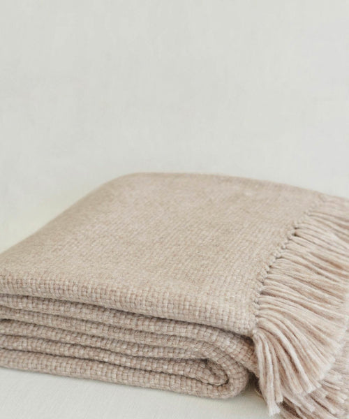 The Large Alpaca Basketweave Throw, featuring a soft woven beige texture and fringe detail on one edge, is neatly folded against a light neutral background.