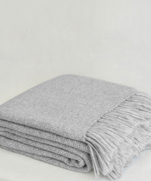The Large Alpaca Basketweave Throw, in light gray with fringed edges and handwoven in Peru, is neatly folded on a plain, light-colored surface.
