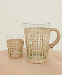 The Seagrass Picnic Pitcher, wrapped in woven wicker, rests on light wood against an off-white background—ideal for an elegant woven tabletop display.