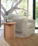 A cozy white armchair with a gray fringed blanket sits by a window. A light wood side table holds a vase of flowers and a candle. Through the glass, a large tree is visible, and the Sedona Jute Rug adds texture to the floor.