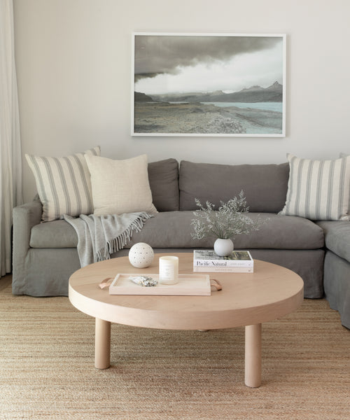 A cozy living room features a gray sofa with striped pillows, a light wood coffee table holding decor, and books. Above the sofa hangs a landscape photo, while the Sedona Jute Rug adds warmth and texture to the floor.