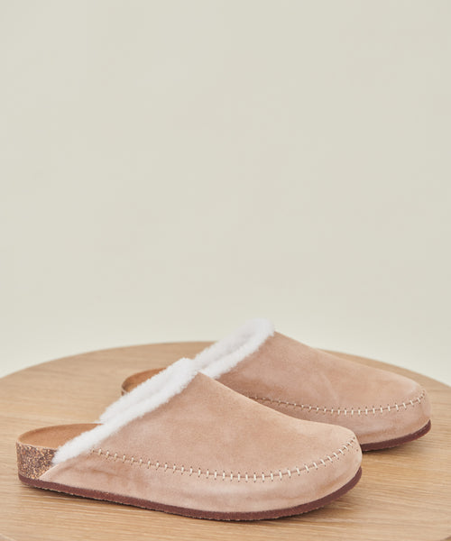 The Shearling-Lined Moc Clog is a tan suede slip-on with a cork sole and white faux fur lining, visible stitching, shown on a round wooden surface against a plain light background.