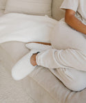 A person in cozy cream loungewear and Shearling Moroccan Slippers with suede outsoles sits cross-legged on a beige couch, next to a textured white blanket.
