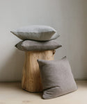 The Sonoma Pillow, a cozy gray cushion, is styled with two additional gray cushions on and beside a natural wooden stool against a light wall and floor, creating a minimalist and welcoming look.
