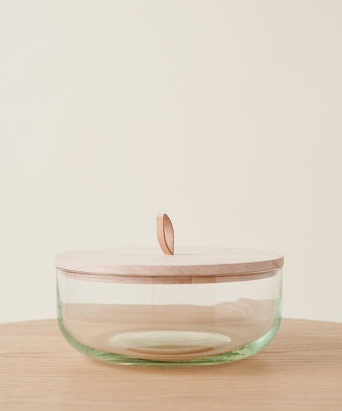 The Stowe Eco Glass Bowl, made from clear recycled glass with a light FSC-certified wooden lid and leather loop handle, rests on a light wood surface against a neutral background.