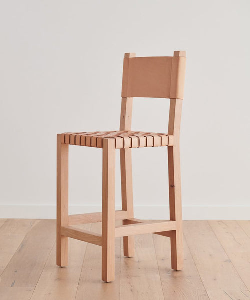 The Tahoe Counter Stool, featuring a woven leather seat and matching backrest on knotty alder wood, stands tall on a light wood floor against a plain white wall.