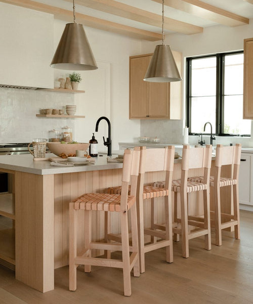 A modern kitchen features light wood Knotty Alder cabinets, a white island with three Tahoe Counter Stools, two metal pendant lights, open shelves displaying dishes, and a black faucet by the window.