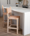 Two Tahoe Counter Stools with woven leather seats are placed by a white kitchen island with a marble countertop. The kitchen features light wood flooring and white cabinetry.