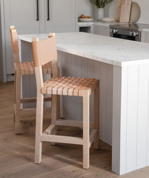 Two Tahoe Counter Stools with woven leather seats are placed by a white kitchen island with a marble countertop. The kitchen features light wood flooring and white cabinetry.