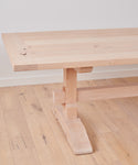 A close-up of the Trestle Dining Table made from knotty alder, showcasing its sturdy leg and thick rectangular top, set on a matching wooden floor against a plain white wall.