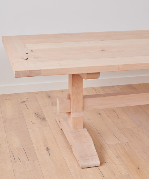 A close-up of the Trestle Dining Table made from knotty alder, showcasing its sturdy leg and thick rectangular top, set on a matching wooden floor against a plain white wall.