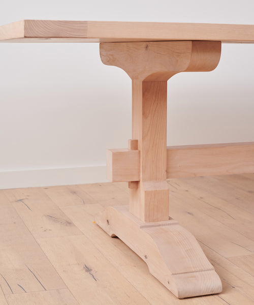 A close-up of the Trestle Dining Table showcases its sturdy handcrafted base and joinery, set on a matching wood floor against a white wall.
