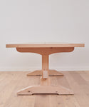 The Trestle Dining Table, handcrafted from Knotty Alder with a central pedestal base, is placed on a light wood floor against a plain white wall and viewed directly from one end.