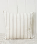 The Ojai Pillow, featuring beige and light gray vertical stripes, is handwoven and rests against a white paneled wall on a light, textured carpet.