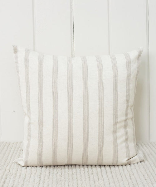 The Ojai Pillow, featuring beige and light gray vertical stripes, is handwoven and rests against a white paneled wall on a light, textured carpet.