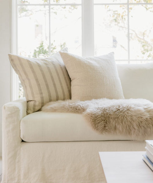 A cozy white sofa with the Ojai Pillow and a beige faux fur throw sits by a large window, bathed in soft natural light. Two neutral pillows and a wooden table with books complete this inviting scene.