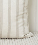A close-up of the Ojai Pillow, featuring a cream color with vertical beige stripes, resting on a ribbed, light fabric surface.