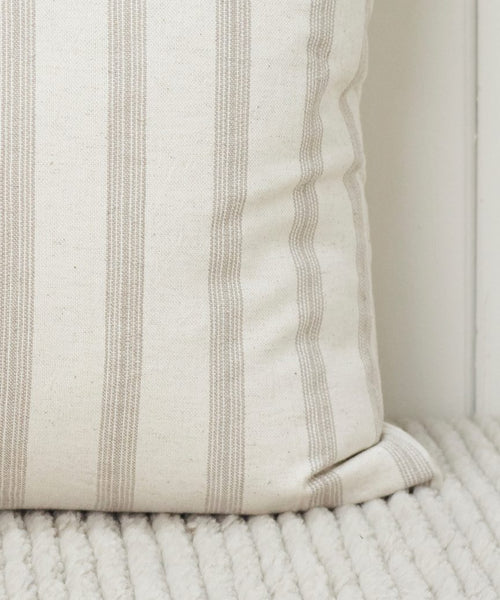 A close-up of the Ojai Pillow, featuring a cream color with vertical beige stripes, resting on a ribbed, light fabric surface.