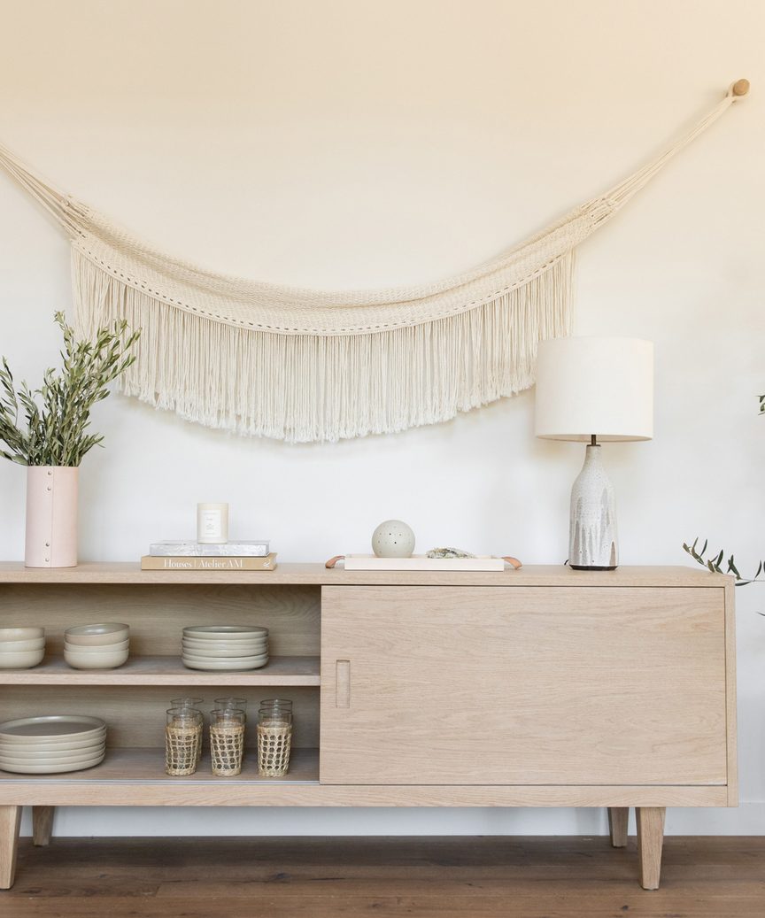 A light wooden sideboard holds stacked plates, glasses, and decorative items. On top are books, a lamp, a vase with greenery, and ceramic decor. Above, the white wall features the Woven Wall Hanging in soft cream tones.