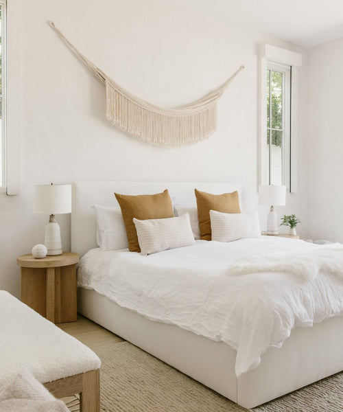 A bright, minimal bedroom features a white bed with tan accent pillows, two bedside tables with lamps, and a Woven Wall Hanging above the bed. Large windows fill the space with natural light, highlighting the handmade decor.
