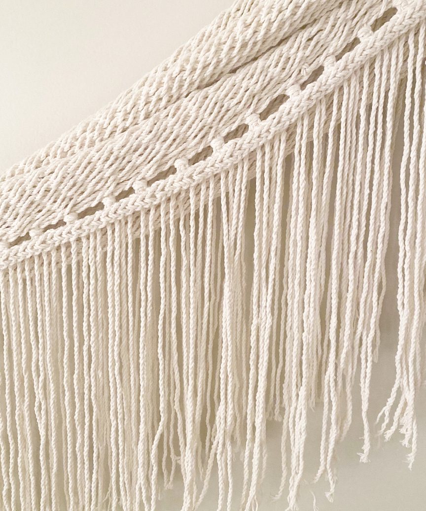 Close-up of the Woven Wall Hanging featuring cream-colored macramé cotton with twisted, knotted rope designs at the top and long, loose fringes cascading below.
