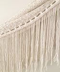 Close-up of the Woven Wall Hanging featuring cream-colored macramé cotton with twisted, knotted rope designs at the top and long, loose fringes cascading below.