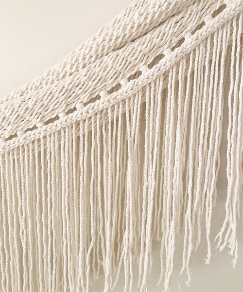 Close-up of the Woven Wall Hanging featuring cream-colored macramé cotton with twisted, knotted rope designs at the top and long, loose fringes cascading below.