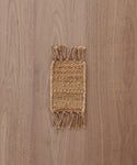 A Rug Swatch, small and rectangular with fringed edges, is displayed on a light brown wooden surface.