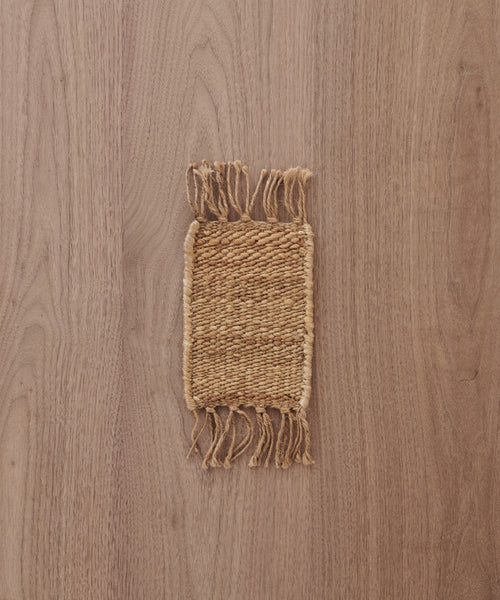 A Rug Swatch, small and rectangular with fringed edges, is displayed on a light brown wooden surface.