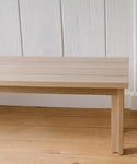 A close-up of the Dining Bench shows its light wood slatted top, placed on a matching floor with a white paneled wall in the background.