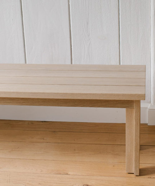A close-up of the Dining Bench shows its light wood slatted top, placed on a matching floor with a white paneled wall in the background.