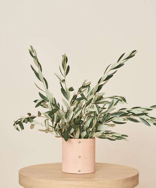 A minimalist arrangement of olive branches in the Small Leather Rivet Vase sits on a round wooden table, the handmade piece’s light pink cylindrical form set against a plain beige background.