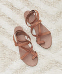 The Oiled Leather Strappy Sandals with ankle straps in brown are displayed side by side on a cream and beige textured rug.