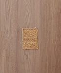 A Rug Swatch, small and square with a woven straw design reminiscent of Sedona rugs, sits on a light brown wooden surface with visible grain patterns.