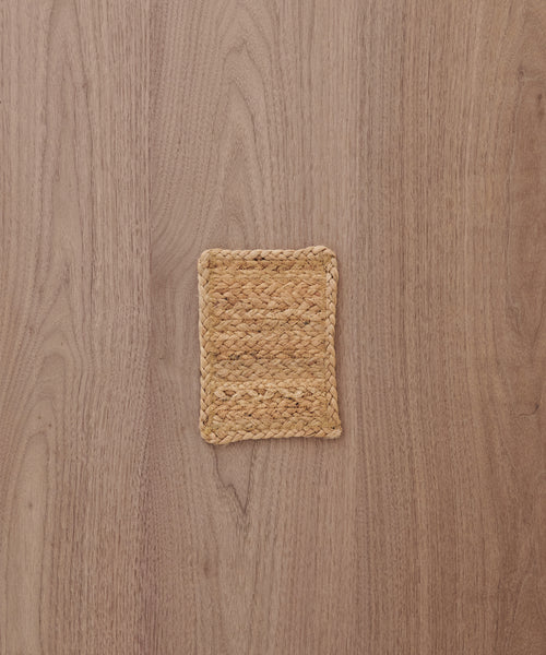 A Rug Swatch, small and square with a woven straw design reminiscent of Sedona rugs, sits on a light brown wooden surface with visible grain patterns.