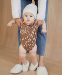 A baby in Shearling Baby Booties, a white knit hat, and a brown onesie with white leaf patterns is held by an adult in blue jeans against a light wood background for a warm, inviting scene.