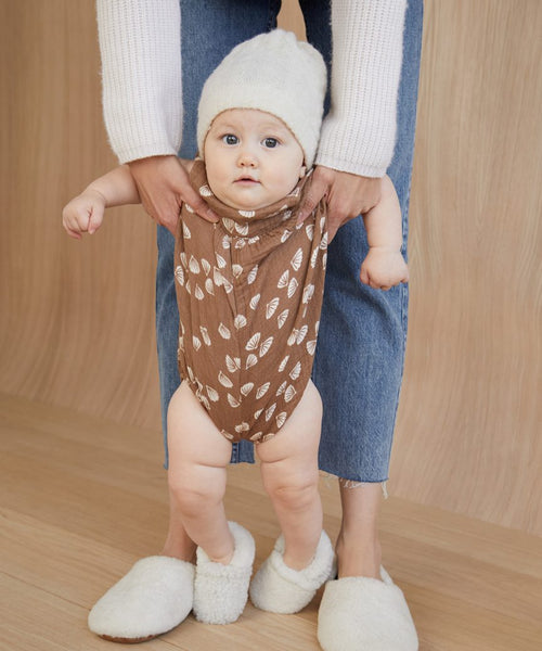 A baby in Shearling Baby Booties, a white knit hat, and a brown onesie with white leaf patterns is held by an adult in blue jeans against a light wood background for a warm, inviting scene.