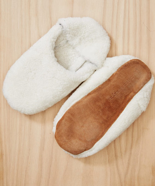 A pair of Shearling Moroccan Slippers—white and fluffy with soft fleece interiors—shown on a wooden surface: one upright displaying the opening, the other flipped to reveal its brown suede outsole.