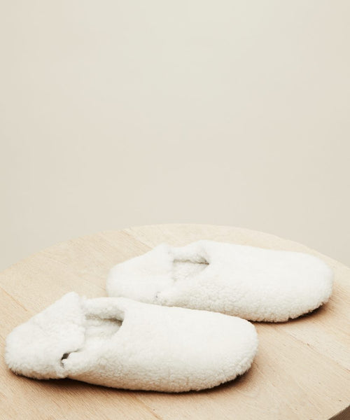 A pair of Shearling Moroccan Slippers with fluffy white uppers and soft suede outsoles sits on light wood against a plain beige background.