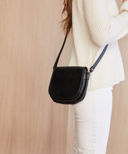 A person wearing a white sweater and pants carries the Small Leather Saddle Bag over their shoulder, standing against a light wooden background.