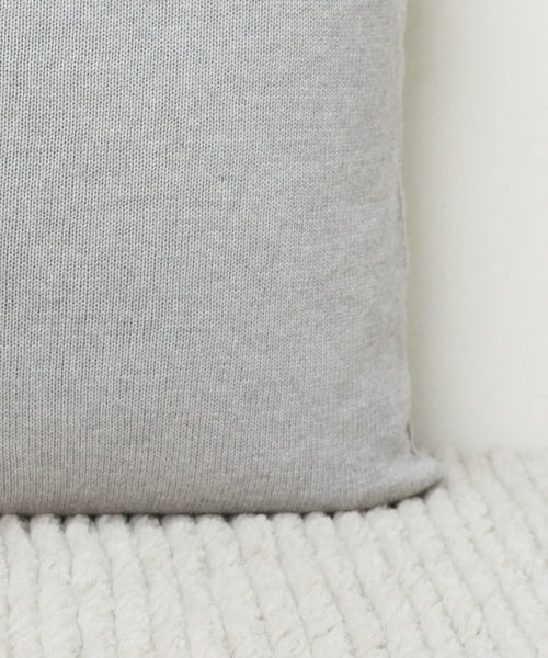 A close-up of the Sonoma Pillow in light gray fabric rests on a white textured surface, with a plain white wall in the background.