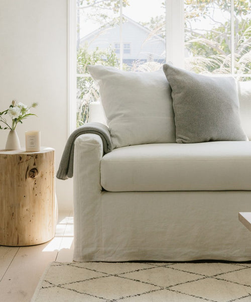 A bright living room features a white sofa with a soft Sonoma Pillow and blanket, plus a wooden stump side table with a vase of flowers and candle. Large windows fill the space with light, while a patterned rug covers the floor.