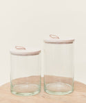 Two Stowe Eco Glass Canisters with light wooden lids and small loop handles sit side by side on a light wood surface, offering stylish, sustainable storage against an off-white background.