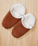 A pair of Suede Moroccan Slippers with brown suede and white fluffy shearling lining, displayed side by side on a light wooden surface.